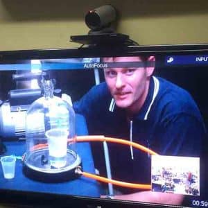 A television screen showing a distance educator running science experiment with a bell jar, vacuum pump and a cup of water. There is an inset of a remote class on the screen and a video conference camera on top of the television.