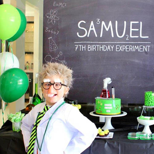 A child wear ing a lab coat and funny glasses whilst standing in front of a blackboard with science motifs and surrounded by green balloons