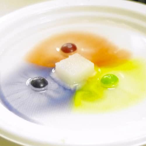 Sugar dissolving in the middle of a white plate, surrounded by 3 dissolving skittles that are forming a cross-shaped pattern