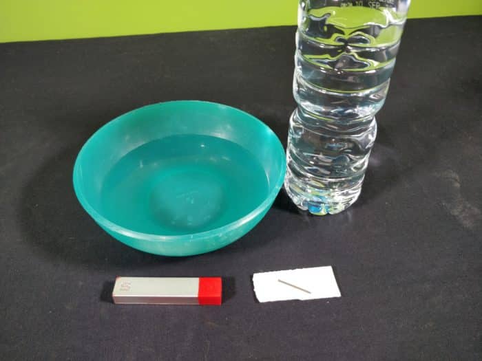 Materials needed to make a simple compass showing a green bowl of water, a magnet, a foam with a needle on top and a bottle of water