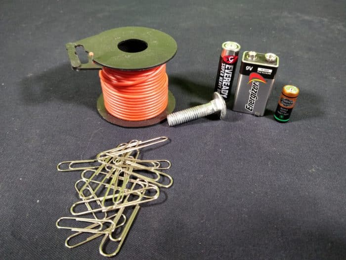 Materials needed to make an electromagnet showing a roll of insulated red wire, paper clips, a steel bolt and a variety of battery sizes