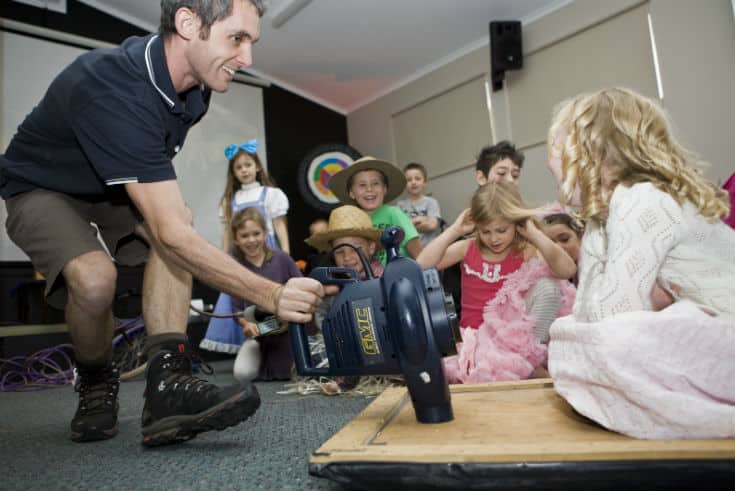 A child sitting on a homemade hovercraft being pushed along by a science presenter