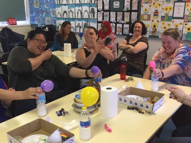 multiple teachers surrounding a table laughing whilst doing an experiment involing a balloon and a bottle