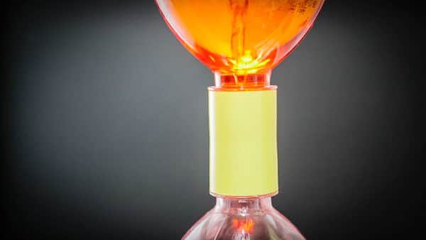 Yellow Vortex valve connecting tow bottles together. Orange liquid is swirling in the top bottle and forming a tornado shaped vortex as it swirls into the bottom bottle