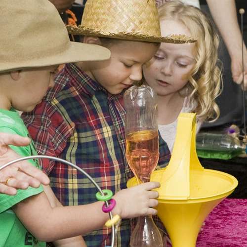 Three children looking at a coin spin down a yellow vortex funnel whilst holding science toys