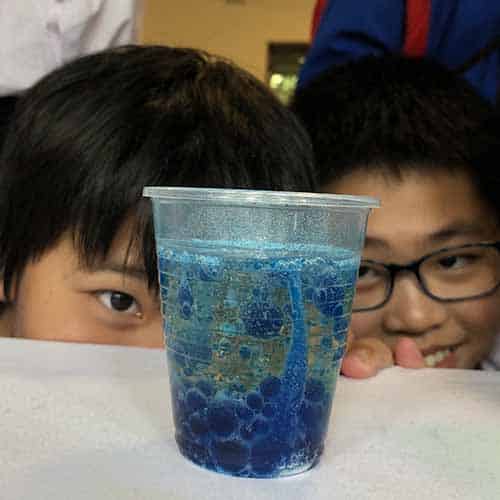 Two smiling students watching a blue lava lamp made in a cup with oil, blue food colouring and alka seltzer