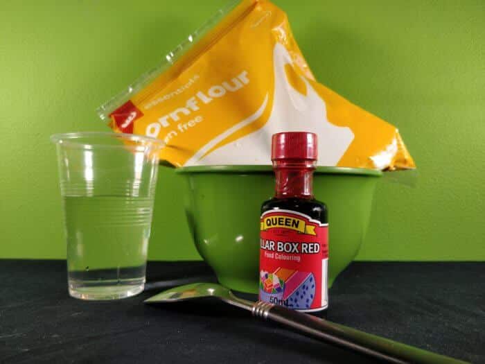 Materials to make corn flour slime showing a cup of water, a bowl, red food colouring and corn flour in a packet