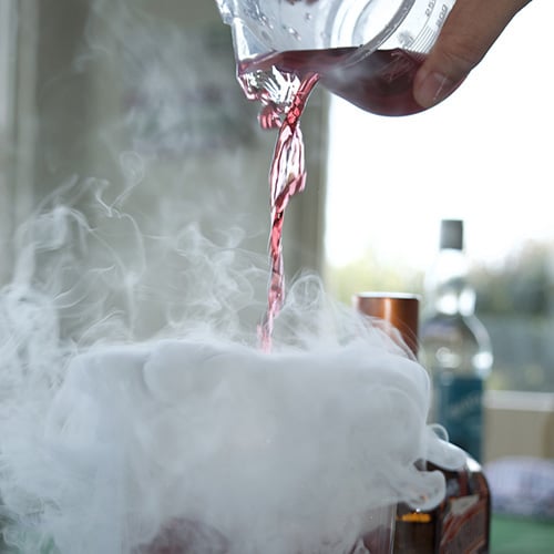 red liquid being poured out of a glass beaker into a a beaker with dry ice fog rushing out and over the table
