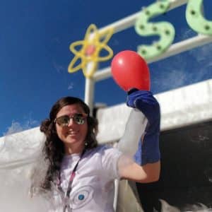 A woman with gloves holding a fogging & iced over soda bottle with a red balloon blown up above it. A science sign on a white tent is in the background