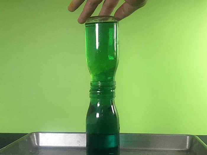 A glass bottle of water suspended over another glass bottle of water. Both bottles are coloured green