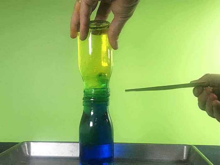 Pulling an index out of between two water bottles that are connected together. (yelloe bottle above and blue bottle below). A swirl of green colour is happening between the two bottles as the blue water below rises into the yellow water above.