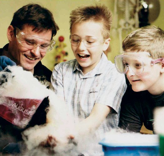 A man pouring dry ice fog out of a beaker whilst two smiling kids watch