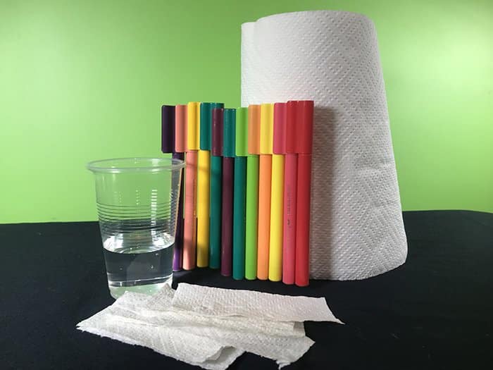 A plastic cup of water, paper towelling, textas an strips of paper towelling on a black desk in front of a green background