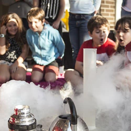 Four kids smiling whilst a fog cloud spills over the side of a science presentation desk past a measuring cylinder, thermos and kettle