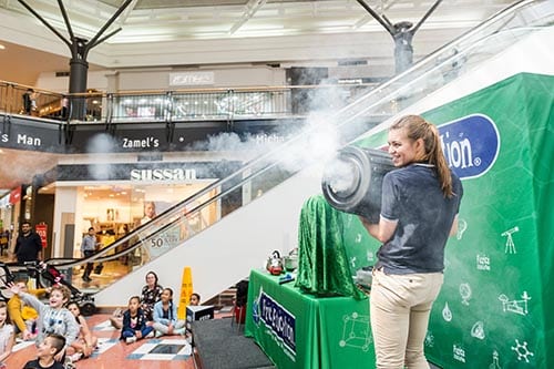 A woman shooting vortex fog rings out of a bin in front of a children;s audience in a shopping centre show