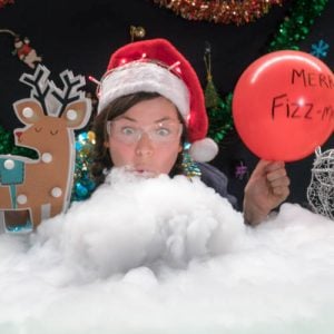 A person wearing a Christmas hat, holding a red balloon, blowing on a bowl of fog next to a reindeer