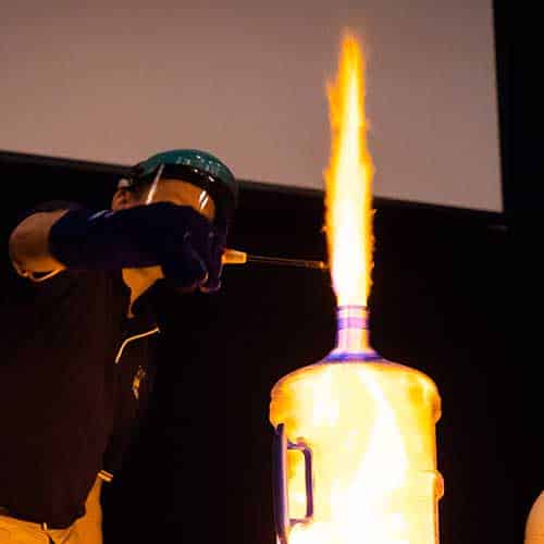 A presenter in full safety gear lighting a gas above a water jug (fire rushing upwards)