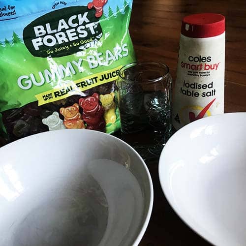 A packet of gummy bears bext two white bowls, a glass of water and a salt container