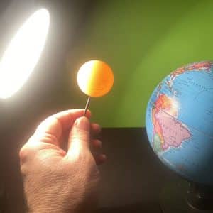 A model of the Earth illuminated by a lamp. A hand is holding a ping pong ball on top of a nail inbetween.