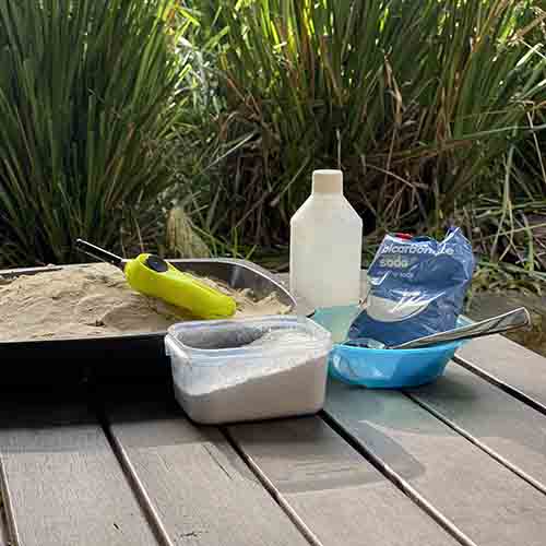 A metal baking tray, a yellow BBQ lighter, a plastic bottle with methylated spirits, a blue bowl and metal spoon, a packet of bicarbonate soda and a plasti container with icing mixture in it