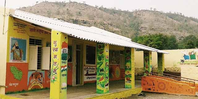 A colourfully painted yellow school building with Indian flag murals & writing as well as paintings of tress, food and people on it