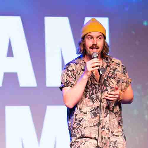 Luke Stellar wearing an orange beanie and a Hawaiian shirt speaking on stage into a microphone 