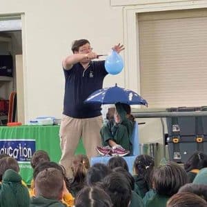 A science presenter holding a water balloon with a flame over a child holding an umbrella