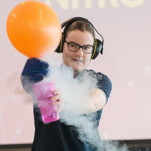 A woman with glasses, safety gloves and ear muffs holding a purple cup with a cloud of water vapour around it and a massive expanding orange balloon