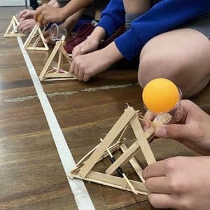 A row of wooden pyramid catapults with ping pong balls ready to fire across a school hall with students